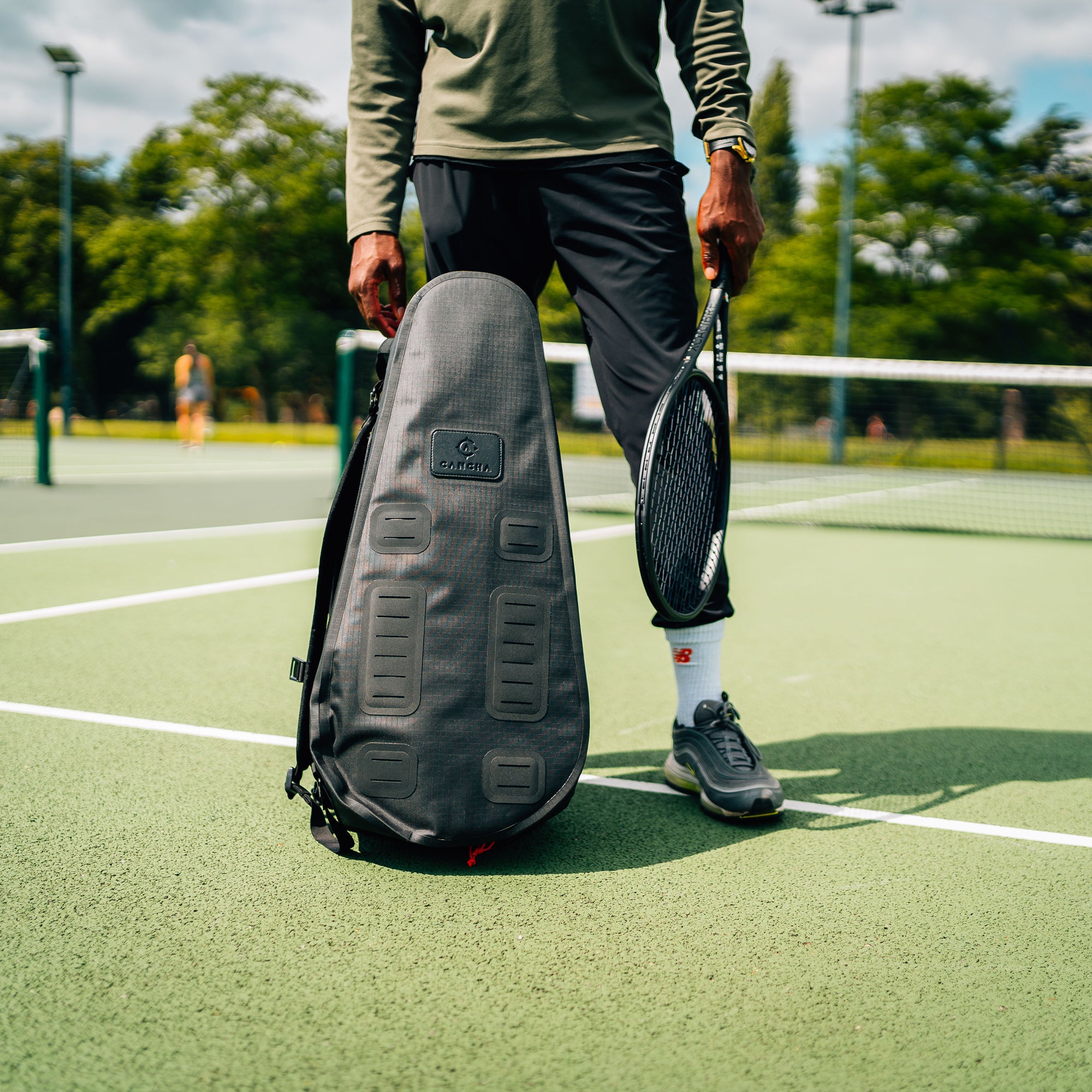 A person carrying a tennis racket in their left hand and a black Cancha Racquet Bag Pro in their right hand stands on an outdoor tennis court. Another player is visible in the background, demonstrating the lightweight performance of their gear.
